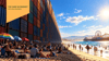A crowded beach beside a towering wall of stacked shipping containers stretching toward the horizon, with the Santa Monica Pier and its Ferris wheel in the distance. A yellow banner in the upper left reads “The Chief Economist with Ryan Severino.”